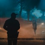 Silhouette of policeman and investigators standing behind crime scene boundary tape at night in forest