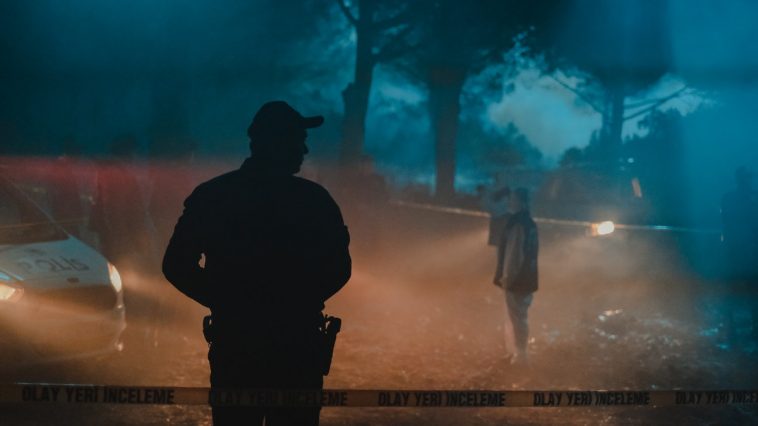 Silhouette of policeman and investigators standing behind crime scene boundary tape at night in forest